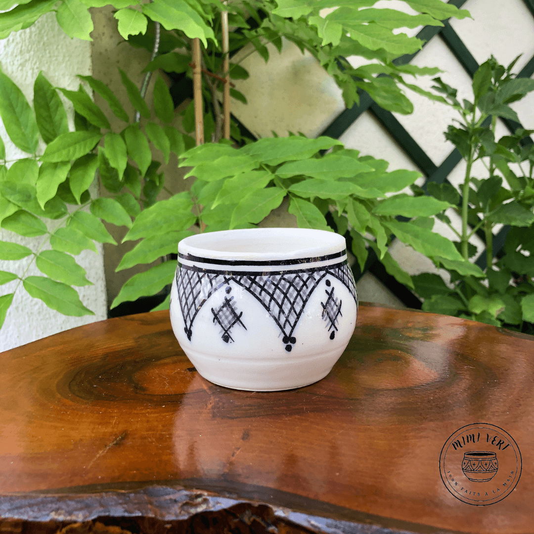 Pot en céramique fait à la main avec des motifs inspirés de l’artisanat marocain, blanc gris et noir, contenant un cactus, posé sur une table en bois avec des plantes vertes en arrière-plan.