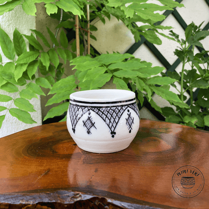 Pot en céramique fait à la main avec des motifs inspirés de l’artisanat marocain, blanc gris et noir, contenant un cactus, posé sur une table en bois avec des plantes vertes en arrière-plan.