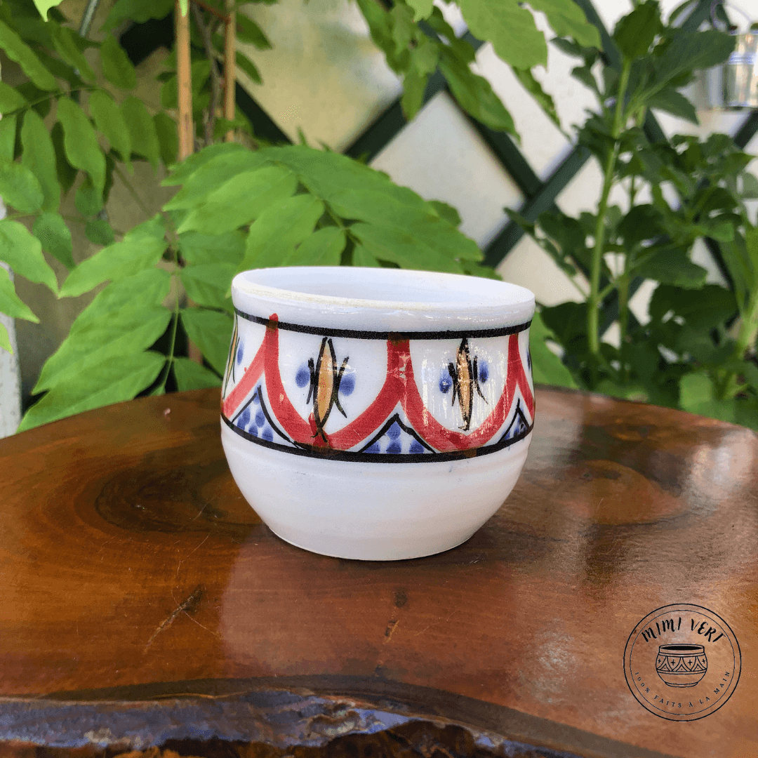 Pot en céramique fait à la main avec des motifs inspirés de l’artisanat marocain, noir et blanc et rouge et jaune, posé sur une table en bois avec des plantes vertes en arrière-plan.