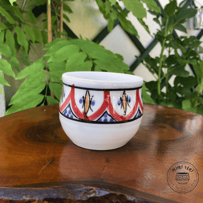 Pot en céramique fait à la main avec des motifs inspirés de l’artisanat marocain, noir et blanc et rouge et jaune, posé sur une table en bois avec des plantes vertes en arrière-plan.