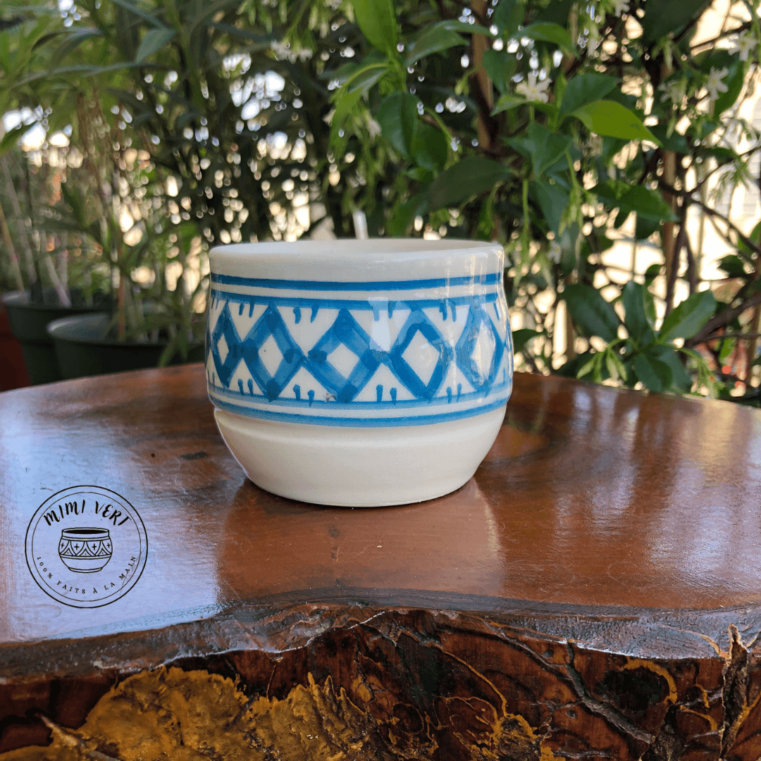 Pot en céramique fait à la main avec des motifs inspirés de l’artisanat marocain, bleu et blanc, posé sur une table en bois avec des plantes vertes en arrière-plan.