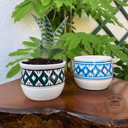 Deux pots en céramique faits à la main avec des motifs inspirés de l’artisanat marocain, posés sur une table en bois avec des plantes vertes en arrière-plan, dont un contenant une plante grasse.