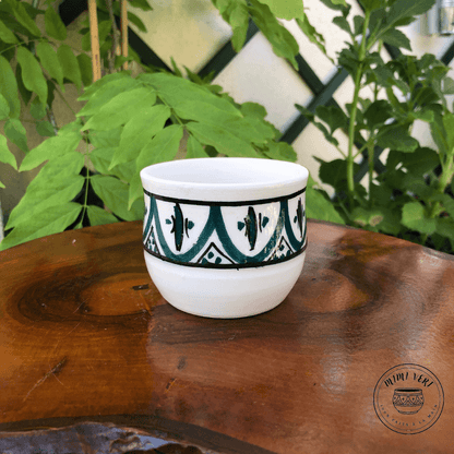 Pot en céramique fait à la main avec des motifs inspirés de l’artisanat marocain, vert noir et blanc, posé sur une table en bois avec des plantes vertes en arrière-plan.