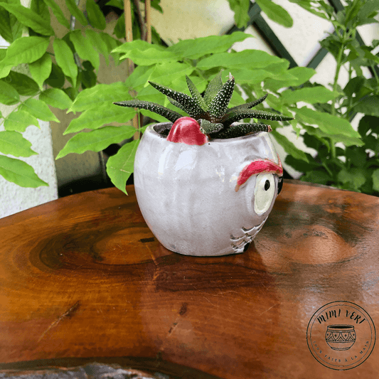 Petit pot en forme de hibou, gris clair, contenant un cactus, posé sur une table en bois avec des plantes vertes en arrière-plan.