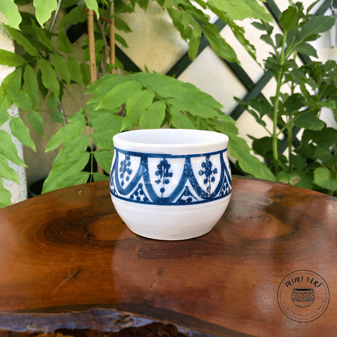 Pot en céramique fait à la main avec des motifs inspirés de l’artisanat marocain, bleu et blanc, posé sur une table en bois avec des plantes vertes en arrière-plan.