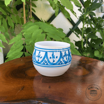 Pot en céramique fait à la main avec des motifs inspirés de l’artisanat marocain, bleu et blanc, posé sur une table en bois avec des plantes vertes en arrière-plan.