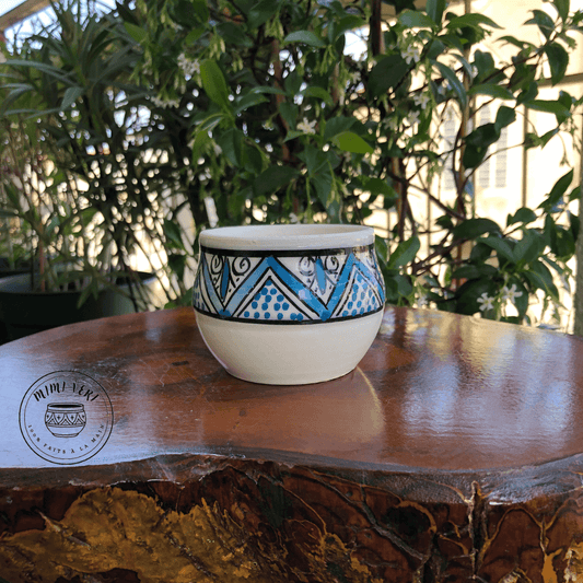 Pot en céramique fait à la main avec des motifs inspirés de l’artisanat marocain, bleu blanc et noir, posé sur une table en bois avec des plantes vertes en arrière-plan.