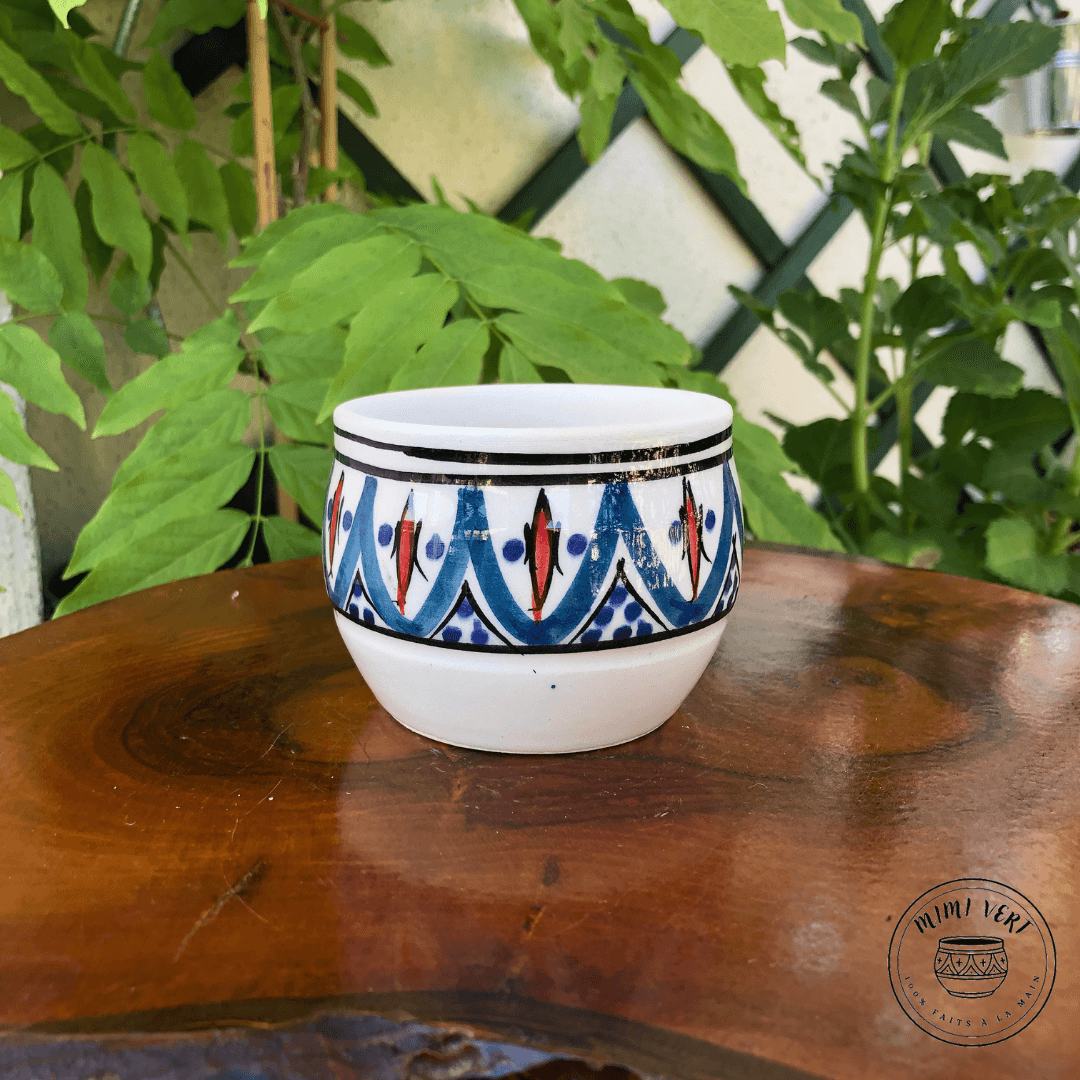 Pot en céramique fait à la main avec des motifs inspirés de l’artisanat marocain, bleu noir et blanc et rouge, posé sur une table en bois avec des plantes vertes en arrière-plan.