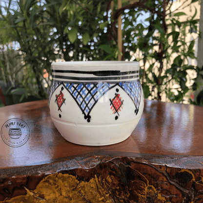 Pot en céramique fait à la main avec des motifs inspirés de l’artisanat marocain, blanc noir rouge et bleu, posé sur une table en bois avec des plantes vertes en arrière-plan.