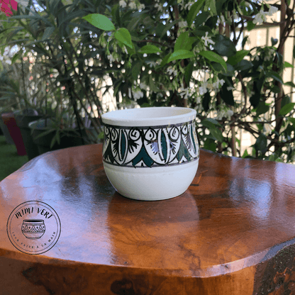 Pot en céramique fait à la main avec des motifs inspirés de l’artisanat marocain, blanc noir et vert, posé sur une table en bois avec des plantes vertes en arrière-plan.