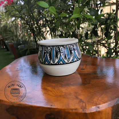 Pot en céramique fait à la main avec des motifs inspirés de l’artisanat marocain, bleu noir et blanc, posé sur une table en bois avec des plantes vertes en arrière-plan.