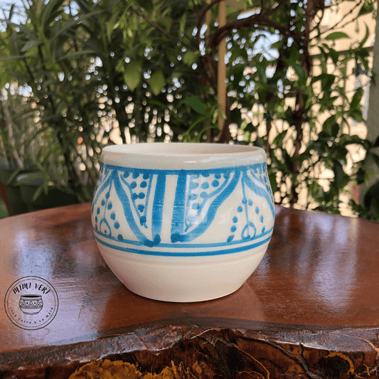 Pot en céramique fait à la main avec des motifs inspirés de l’artisanat marocain, bleu et blanc, posé sur une table en bois avec des plantes vertes en arrière-plan.
