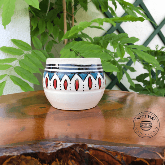 Pot en céramique fait à la main avec des motifs inspirés de l’artisanat marocain, bleu rouge et blanc, contenant une plante grasse, posé sur une table en bois avec des plantes vertes en arrière-plan.