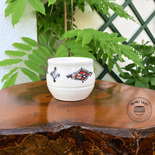 Pot en céramique fait à la main avec des motifs inspirés de l’artisanat marocain, blanc bleu et rouge, posé sur une table en bois avec des plantes vertes en arrière-plan.