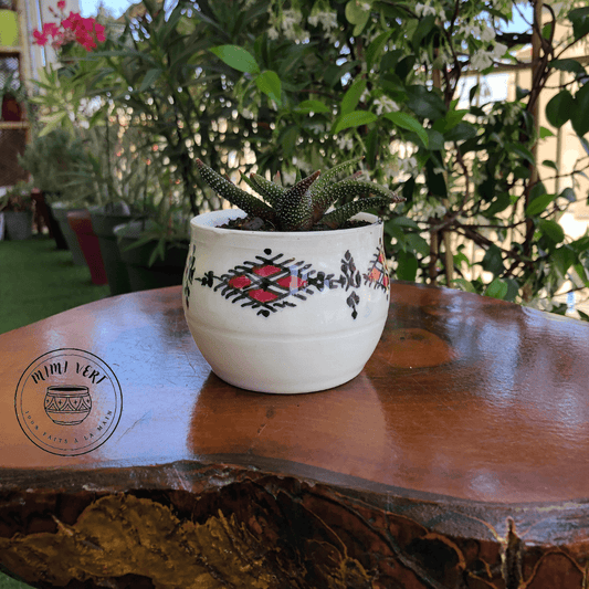 Pot en céramique fait à la main avec des motifs inspirés de l’artisanat marocain, blanc et rouge, contenant une plante grasse, posé sur une table en bois avec des plantes vertes en arrière-plan.