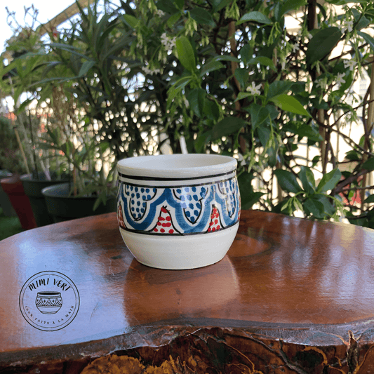 Pot en céramique fait à la main avec des motifs inspirés de l’artisanat marocain, bleu rouge et blanc, posé sur une table en bois avec des plantes vertes en arrière-plan.