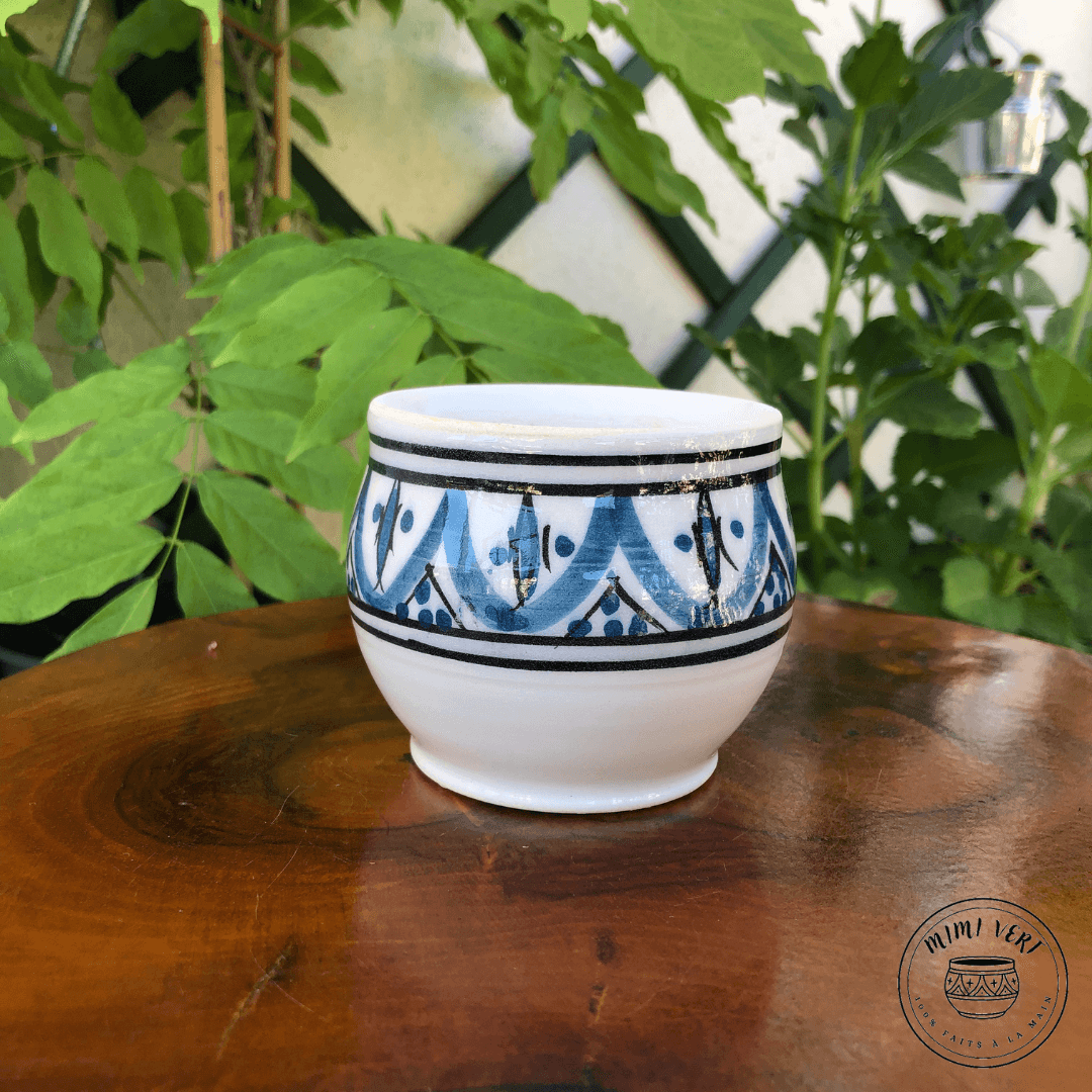 Pot en céramique fait à la main avec des motifs inspirés de l’artisanat marocain, bleu noir et blanc, posé sur une table en bois avec des plantes vertes en arrière-plan.