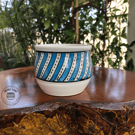 Pot en céramique fait à la main avec des motifs inspirés de l’artisanat marocain, bleu et blanc, posé sur une table en bois avec des plantes vertes en arrière-plan.