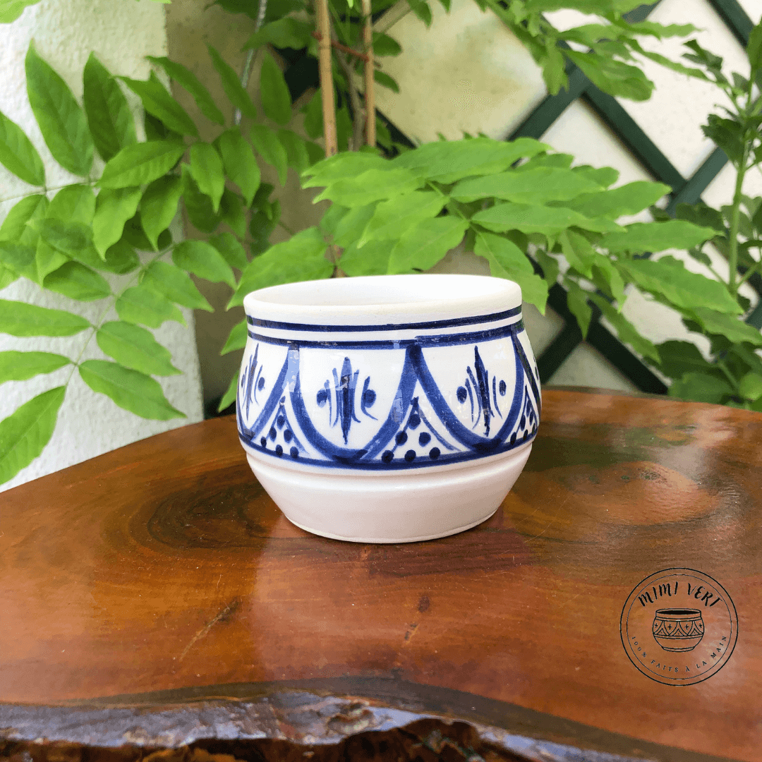 Pot en céramique fait à la main avec des motifs inspirés de l’artisanat marocain, blanc et bleu, posé sur une table en bois avec des plantes vertes en arrière-plan.